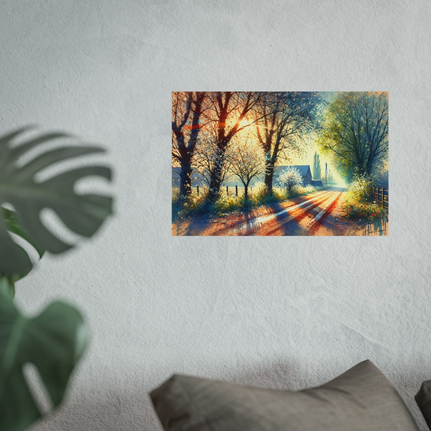 Art print of a sunlit forest path on a wall with a plant and pillow in the foreground.