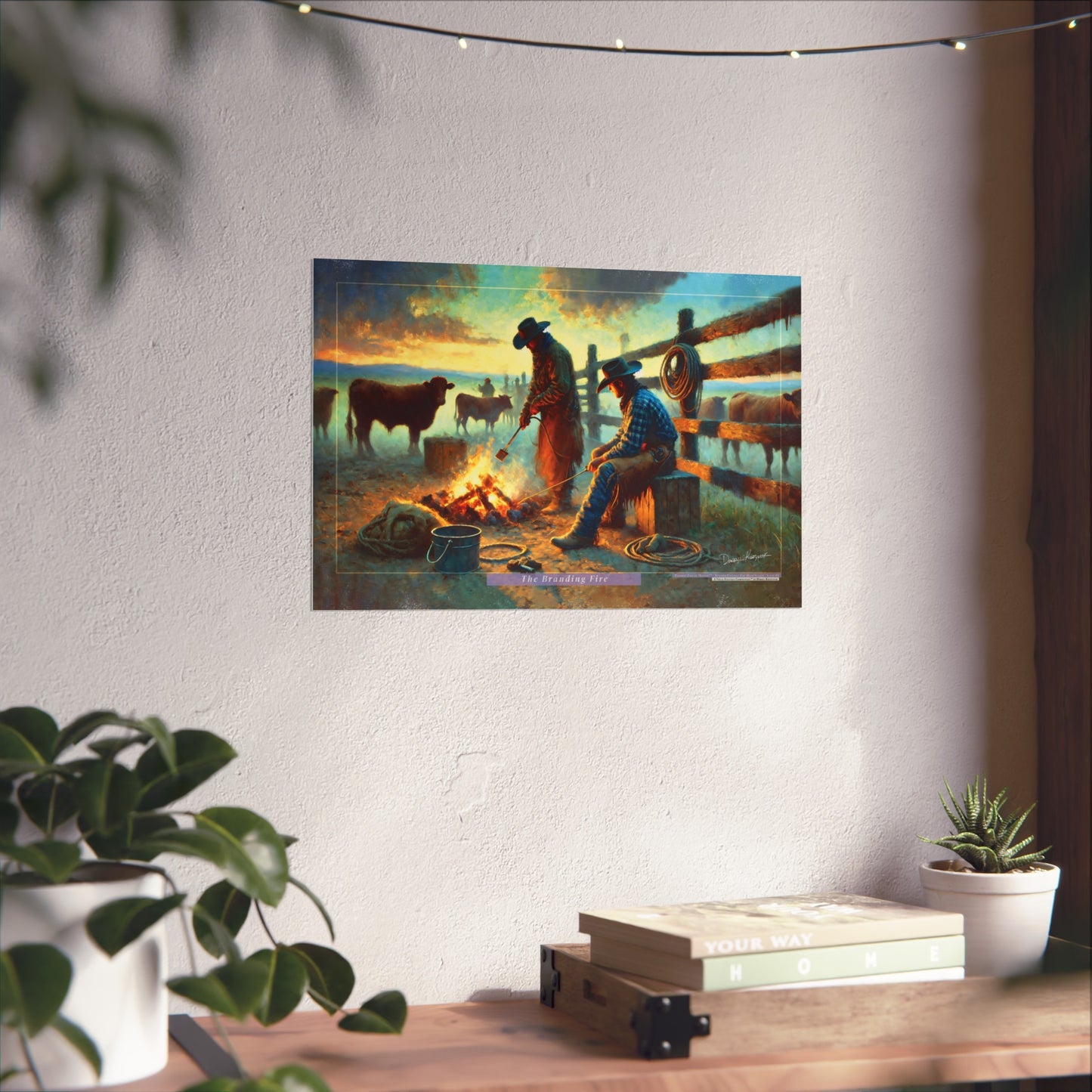 Poster of two cowboys by a branding campfire on a wall with plants and books below.
