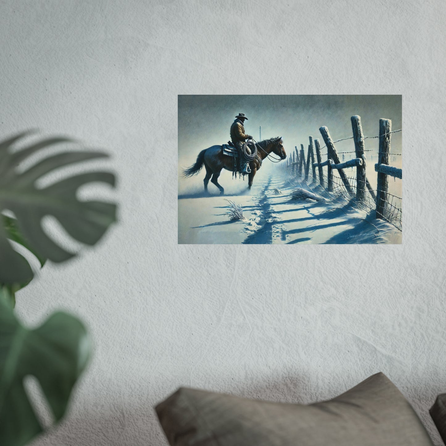 Poster of a cowboy on horseback by a fence on a wall with a plant and pillow in the foreground.