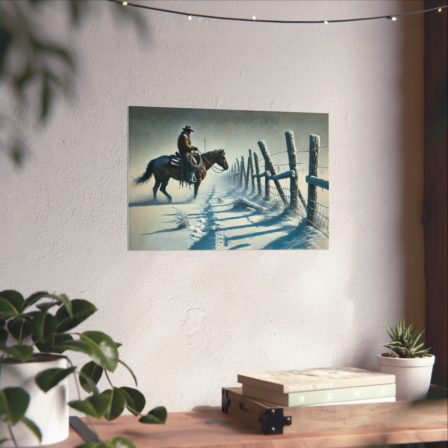 Poster of a cowboy on horseback leaning against a fence on a wall above a desk with plants and books.