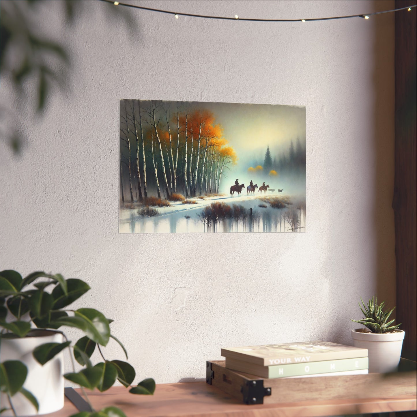 Framed artwork of a winter landscape with trees and cowboys on a wall above a wooden table with plants.