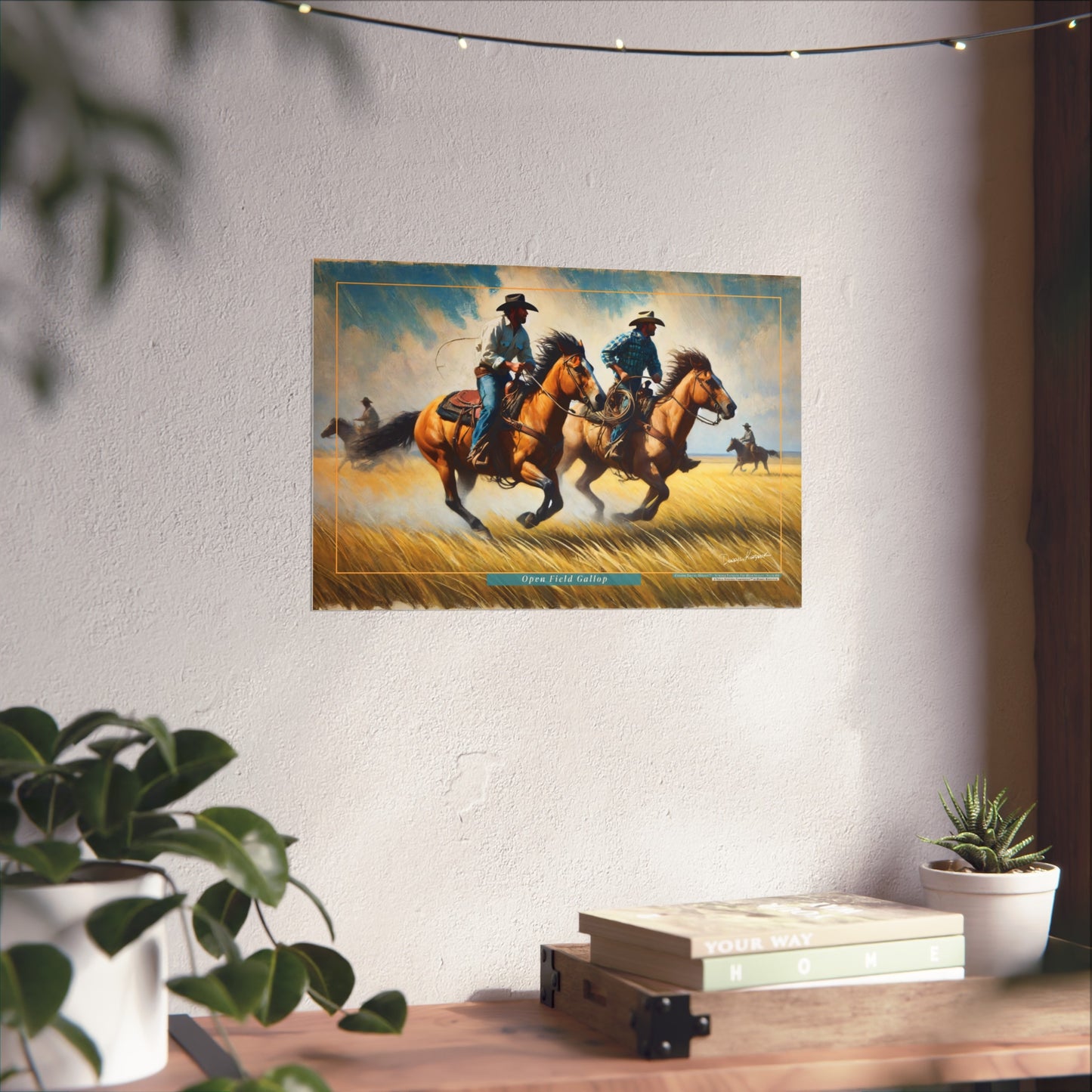Framed artwork of cowboys on horses hanging on a wall above a desk with plants.