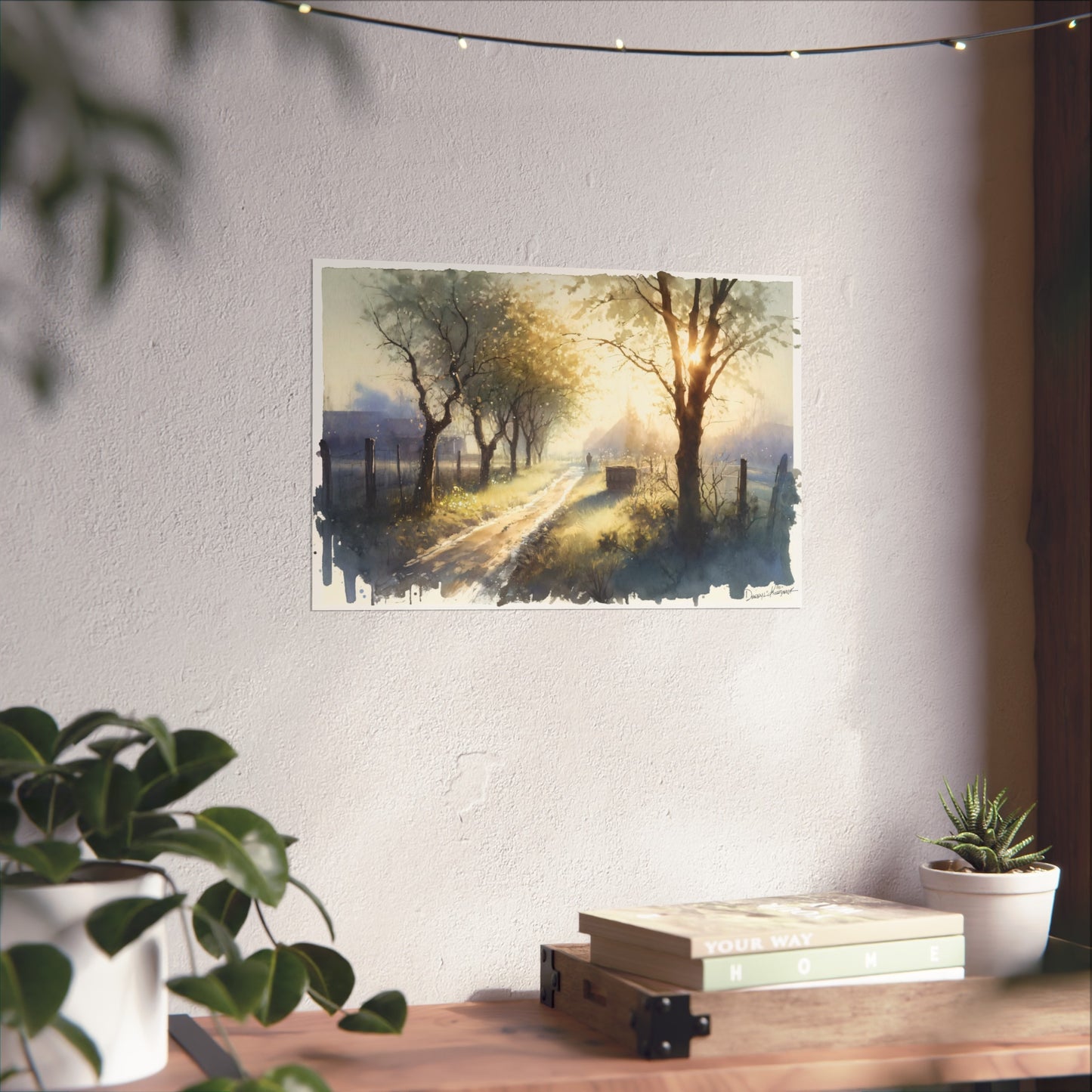 Fine art print of a scenic landscape with trees and a path on a wall above a desk with plants and books.